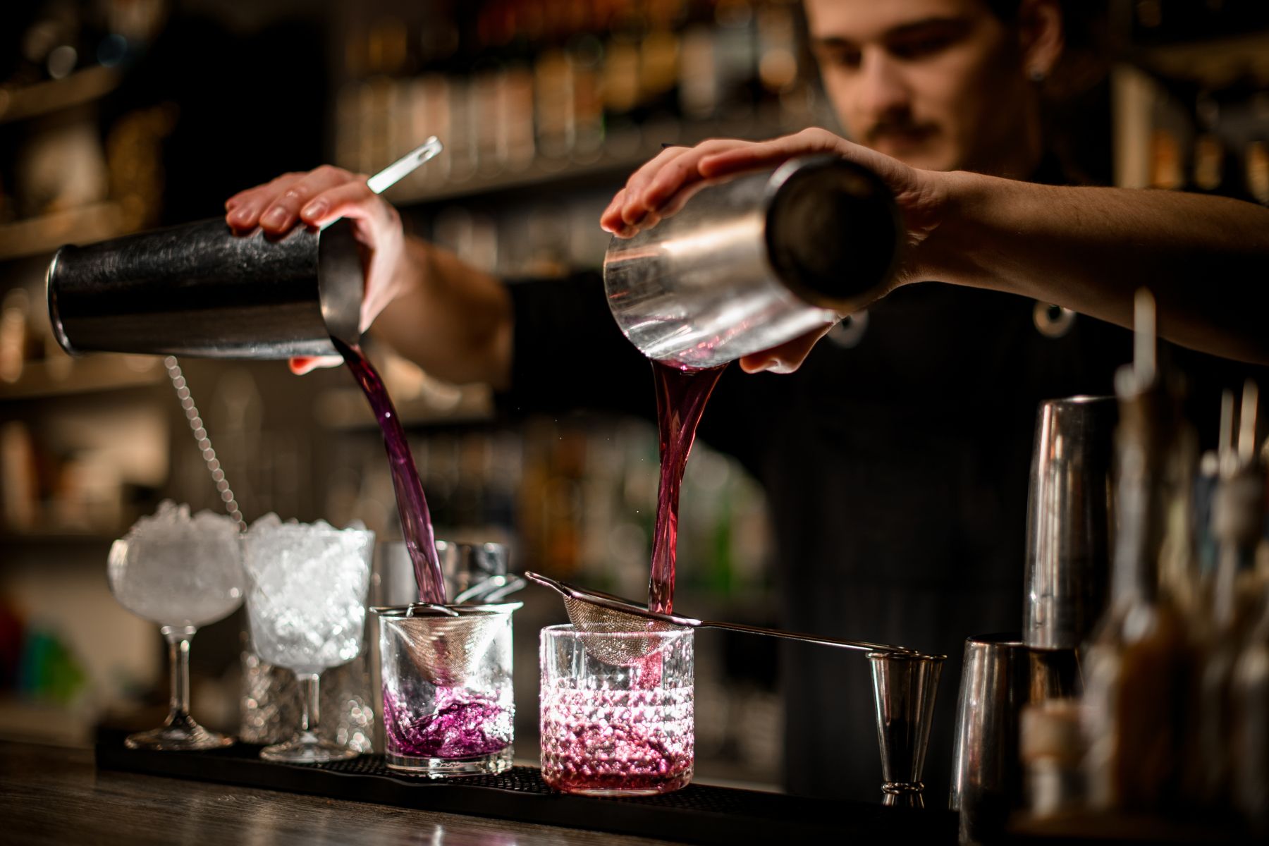 Bartender pours a vibrant purple cocktail from a shaker into ice-filled glasses at a bar.