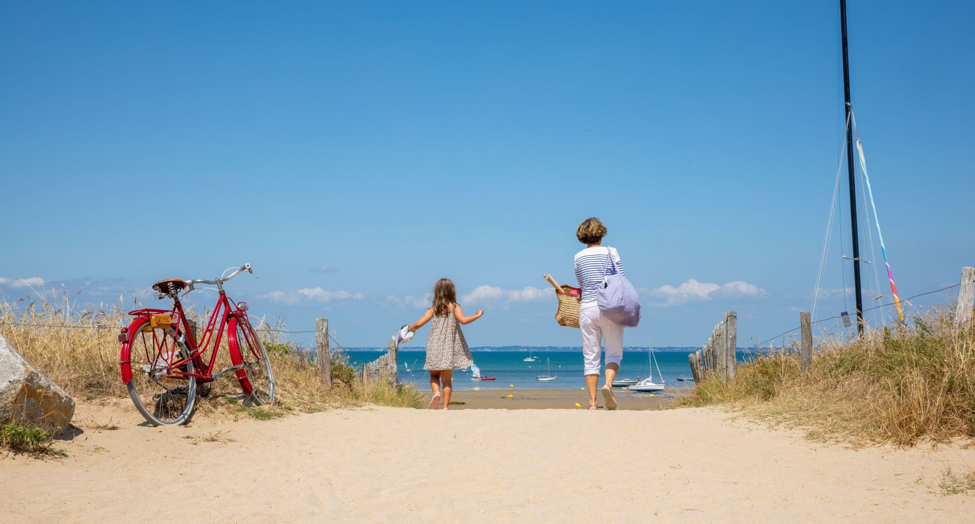 A woman and a child walk toward the sea on a sandy path, carrying bags, with a red bicycle resting on the left and sailboats in the water ahead.