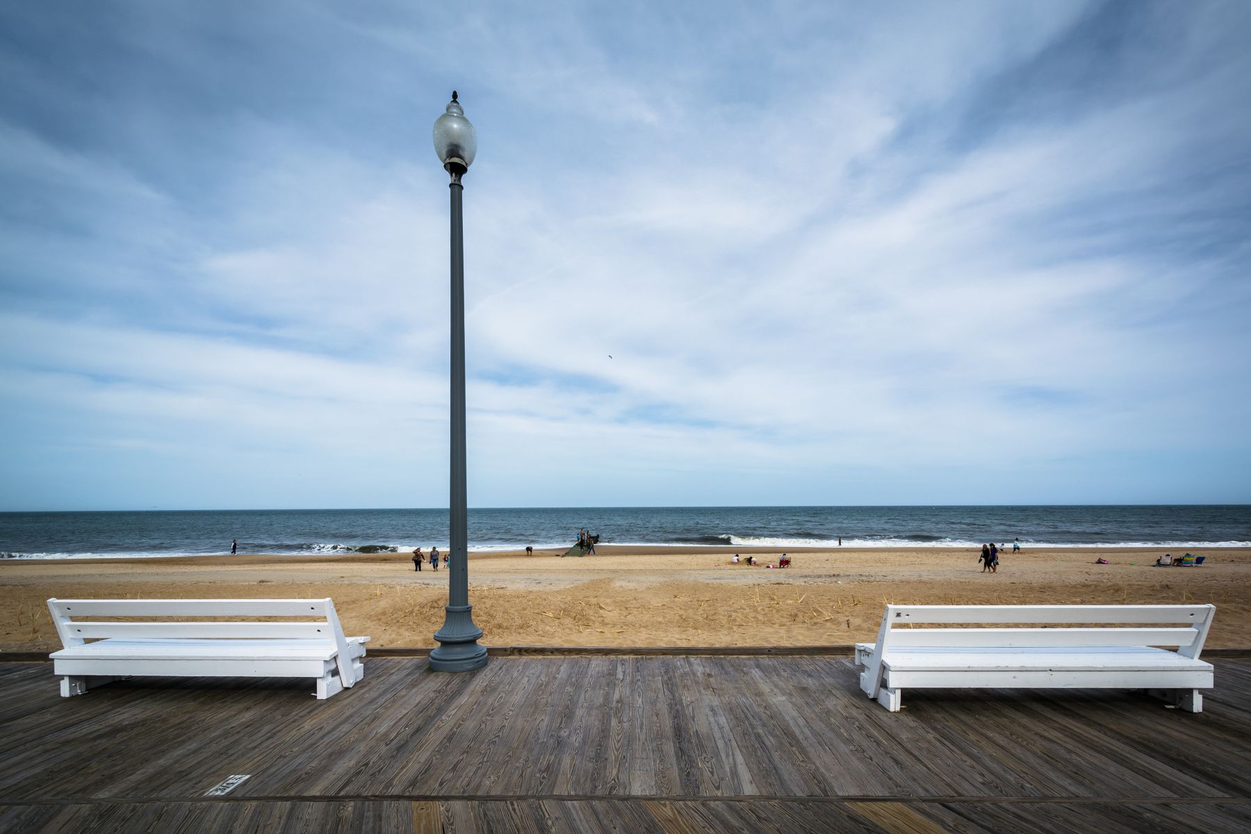 Beach scene with a tall lamppost on a wooden boardwalk, white benches, and people on the sandy shore by the ocean under a blue sky.