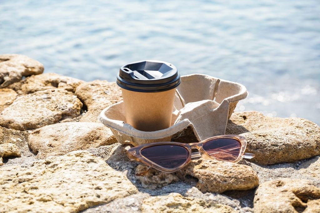 To-go coffee cup with lid on a rocky shore beside calm water, with pink sunglasses nearby.